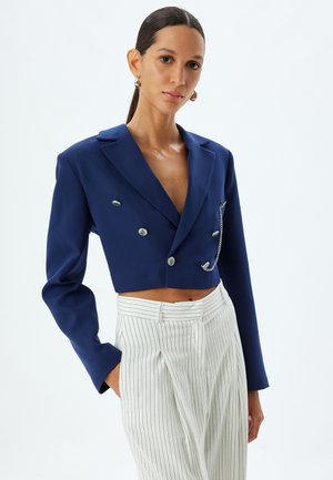 Marineblauwe cropped blazer met dubbele zilveren knopen en kettingdetail op de voorzak, gecombineerd met wit gestreepte broek.