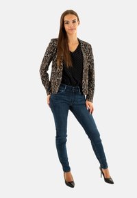 Blazer à motif noir et or sur un top noir à pois, associé à un jean skinny bleu foncé et des escarpins noirs à talon pointu.