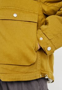 Gele denimjas met een grote borstzak, drukknoopsluiten en een verstelbare trekkoord aan de onderkant. Gladde textuur, robuust ontwerp.