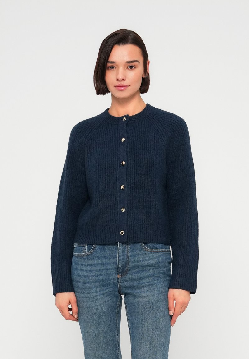 Καρντινάλιος ναυτικού μπλε χρώματος κατασκευασμένος από υφασμά υφαντός με υφή, με στρογγυλή λαιμόκοψη και κλείσιμο μπροστινών κουμπιών με μεταλλικές λεπτομέρειες.