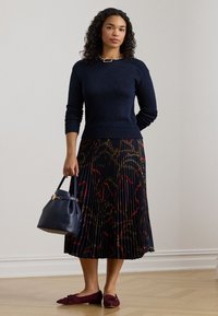 Marineblauer gestrickter Pullover mit geripptem Saum, kombiniert mit einem plissierten schwarzen Rock mit buntem Kettenmuster, dunklen flachen Schuhen und einer marineblauen Tasche.