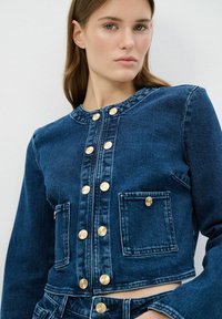 Chaqueta de mezclilla azul oscuro con un diseño cortado, botones dorados, dos bolsillos frontales y un escote redondeado. Textura suave y ajuste estructurado.