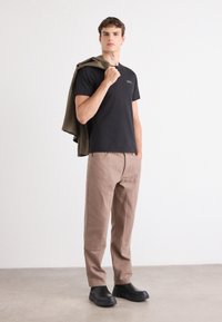 Tricou negru, pantaloni bej cu cusături texturate și pantofi negri. Jachetă verde măslinie purtată pe umăr. Design minimalist.