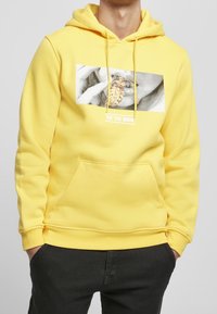 Muž v žluté mikině s přední kapsou, černých kalhotách a grafikou rtů, které drží zlatý pendant s textem "TIC TAC BOOM" pod ním.