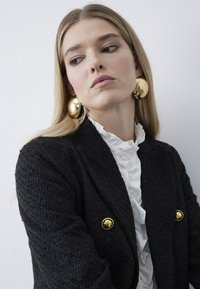 Blond kvinde i struktureret sort blazer med guldknapper og store guldøreringe, iført en hvid bluse med flæser, kigger sidelæns mod en hvid væg.