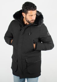 Veste parka noire avec une capuche en fausse fourrure, fermeture éclair et boutons-pression, deux poches avant et des poignets côtelés. Tissu doux et isolant.