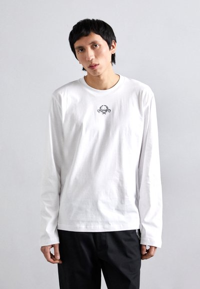 T-shirt blanc à manches longues en coton, avec un petit graphique noir brodé sur la poitrine et un ourlet droit. Porté avec un pantalon noir.