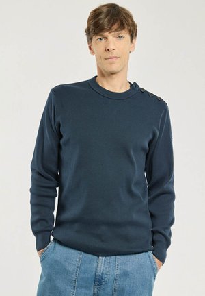 Homme aux cheveux châtain clair portant un pull bleu foncé avec des boutons sur l'épaule et un jean bleu clair, mains dans les poches, fond neutre.