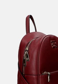 Sac à dos en cuir bordeaux avec zips dorés, partie supérieure arrondie et finition texturée. Il dispose d'un accès à double zip et d'une poignée supérieure pour le transport.