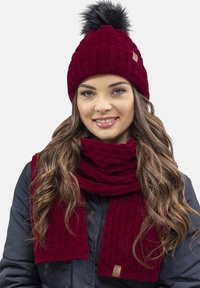 Berretto a maglia bordeaux con pompon nero, abbinato a una sciarpa a coste coordinata. Entrambi gli articoli presentano un motivo testurizzato e un'etichetta di marca.