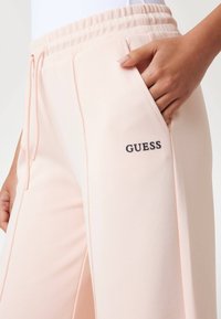 Gaiši rozā sporta bikses ar elastīgu jostasvietu, auklu un sānu kabatām, uz priekšu ir arvien redzams tumši zils "GUESS" logo.