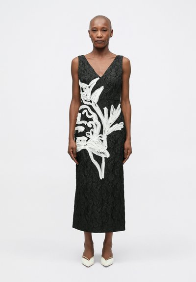 Robe noire avec un tissu texturé, décolleté en V profond, broderie florale blanche d'un côté et coupe droite. Portée avec des escarpins blancs à bout pointu.