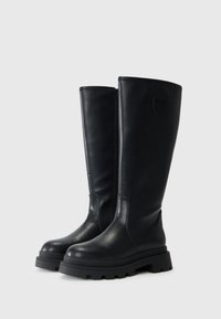 Botas de cuero negras hasta la rodilla con suela de goma gruesa, textura lisa y un acento de logo en el eje superior. Detalle de costura en el lateral.