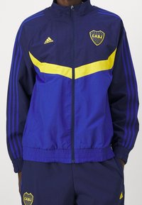 Námořnická modrá sportovní bunda se žlutými akcenty, zipovým zapínáním a designem se třemi pruhy. Obsahuje emblema CABJ na hrudi a elastické manžety.