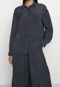 Navy button-up shirt with a textured, crinkled fabric, featuring a chest pocket and long sleeves, paired with wide-leg trousers.