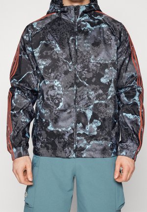 Homme portant une veste zippée à motif camouflage noir et bleu avec des bandes orange sur les manches et un pantalon bleu sarcelle.