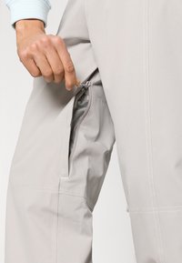 Calças cinza claro com um bolso com fecho éclair, apresentando um forro em rede no interior do bolso e uma textura de tecido suave e durável.