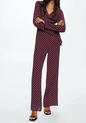 Kvinde stående med armene krydsede iført en vinrød langærmet polkaprikket jumpsuit og sorte åbne stiletter på hvid baggrund.