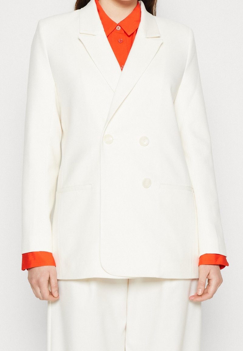 Vit dubbelknäppt blazer med spetsiga slag, sidofickor och röda manschetter, tillverkad av ett slätt, strukturerat tyg.