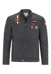 Gray jacket with a zip front, stand collar, and multiple patches, including a flag and logos. Features two side pockets and a textured finish.