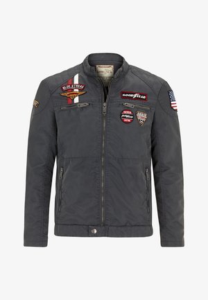 Szürke kabát, cipzáros elülső résszel, álló gallérral és több folttal, beleértve egy zászlót és logókat. Két oldalsó zsebe van és texturált felülettel rendelkezik.