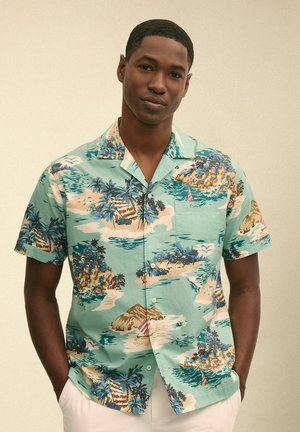 Młody mężczyzna w jasnoniebieskiej koszuli z krótkim rękawem, ozdobionej wzorami tropikalnej wyspy i żaglówki, stojący z rękami w kieszeniach.