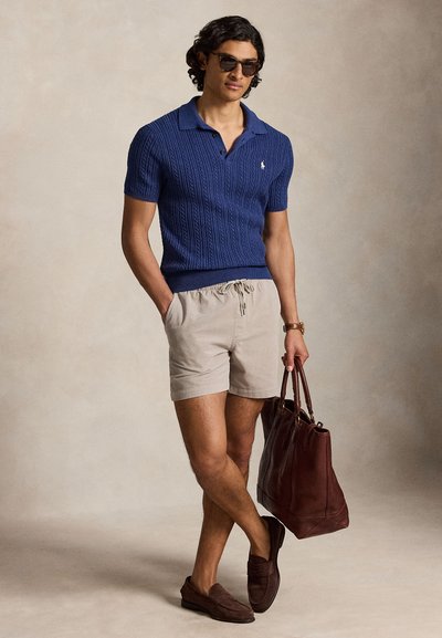 Homme en polo en maille bleu à manches courtes, short beige, mocassins marron et lunettes de soleil, tenant un grand sac cabas en cuir marron foncé.