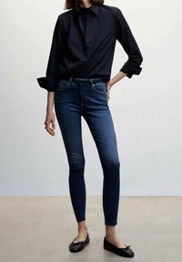 Slim-fit dark blue jeans paired with a black buttoned shirt and black ballet flats on a standing person against a plain background.