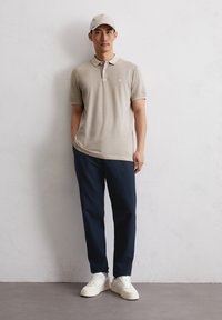 Beżowa koszulka polo z krótkim rękawem i logo na piersi, w połączeniu z granatowymi spodniami i białymi trampkami; neutralny kolor czapki dopełnia stylizację.