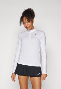 Maglietta sportiva bianca a maniche lunghe con colletto zip e texture a coste, abbinata a una skort nera con logo Nike.