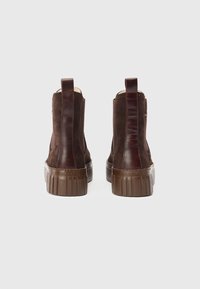 Par de botas de camurça castanhas até ao tornozelo com faixa de couro posterior e solas de borracha grossas, vistas de trás sobre um fundo branco.