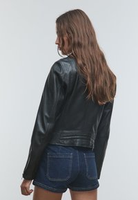 Frau mit langen braunen Haaren, die eine schwarze Lederjacke und dunkelblaue Jeansshorts trägt, seitlich stehend vor einem neutralen Hintergrund.