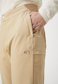 Beiga byxor i ett texturerat tyg, med en sidficka med dragkedja och en logotyp tryckt i en ljusare nyans på låret.