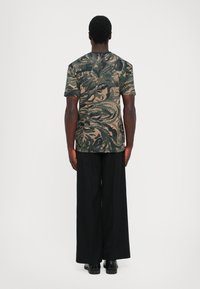 Hombre vestido con una camisa de manga corta de estampado abstracto en verde oscuro y beige, pantalones anchos negros y zapatos negros, de pie de espaldas sobre un fondo blanco.