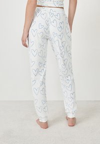 Persoon in witte pyjamabroek met een blauw hartpatroon, staand op blote voeten op een vloer van tapijt, gezien vanaf de achterkant.
