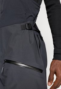 Sötétszürke, sima textúrájú kültéri nadrág, oldalsó cipzáras zsebbel és állítható pánttal a biztos illeszkedés érdekében.