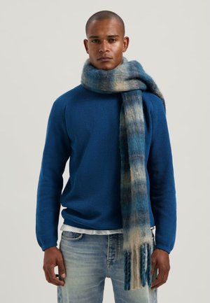 Man in een blauwe gebreide trui en een lange blauwe gradient wol sjaal, gecombineerd met lichtgewassen jeans, staande voor een effen achtergrond.