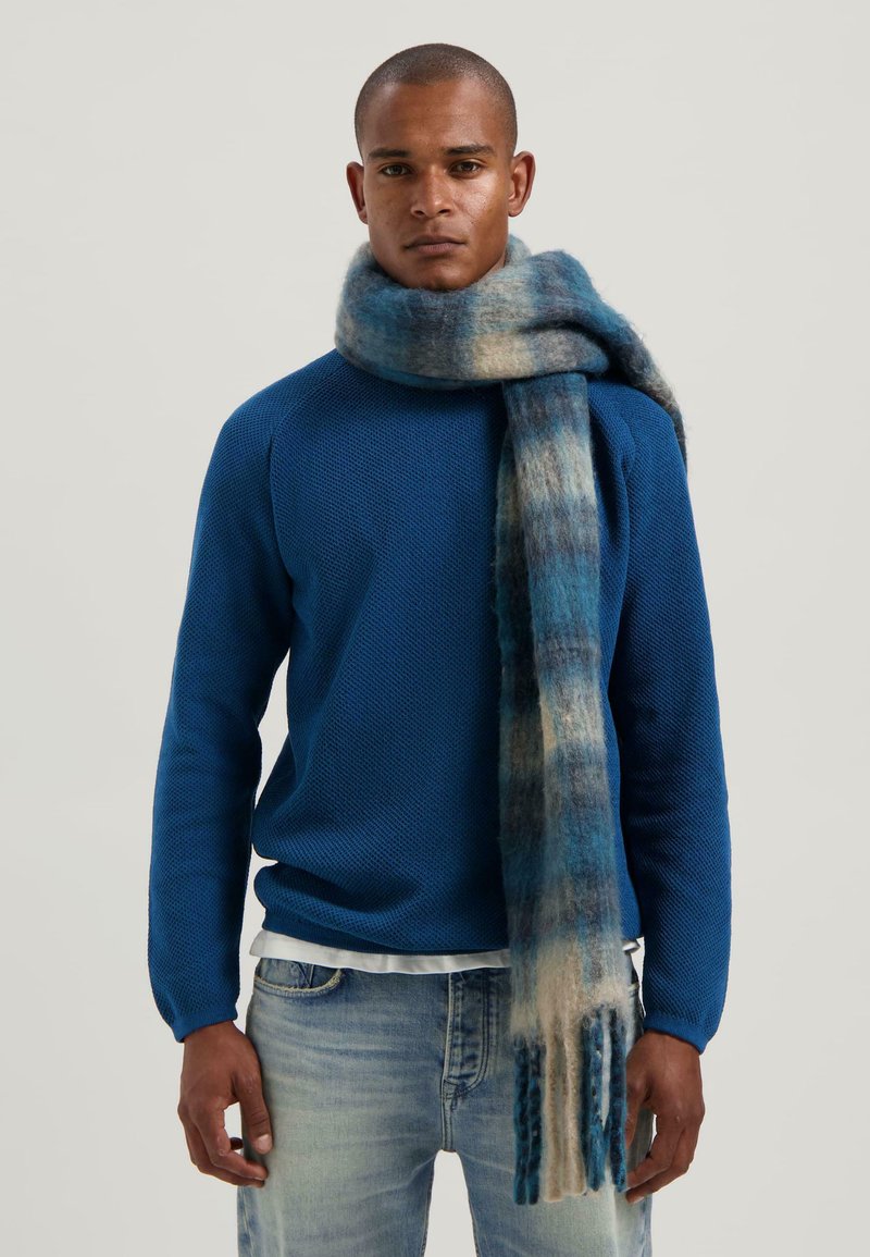 Man in een blauwe gebreide trui en een lange blauwe gradient wol sjaal, gecombineerd met lichtgewassen jeans, staande voor een effen achtergrond.