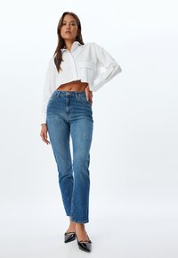 Lyhennetty valkoinen paitapusero pitkillä hihoilla, yhdistettynä keskikorkeisiin sinisiin suoriin farkkuihin ja mustiin teräväkärkisiin ballerinoihin.