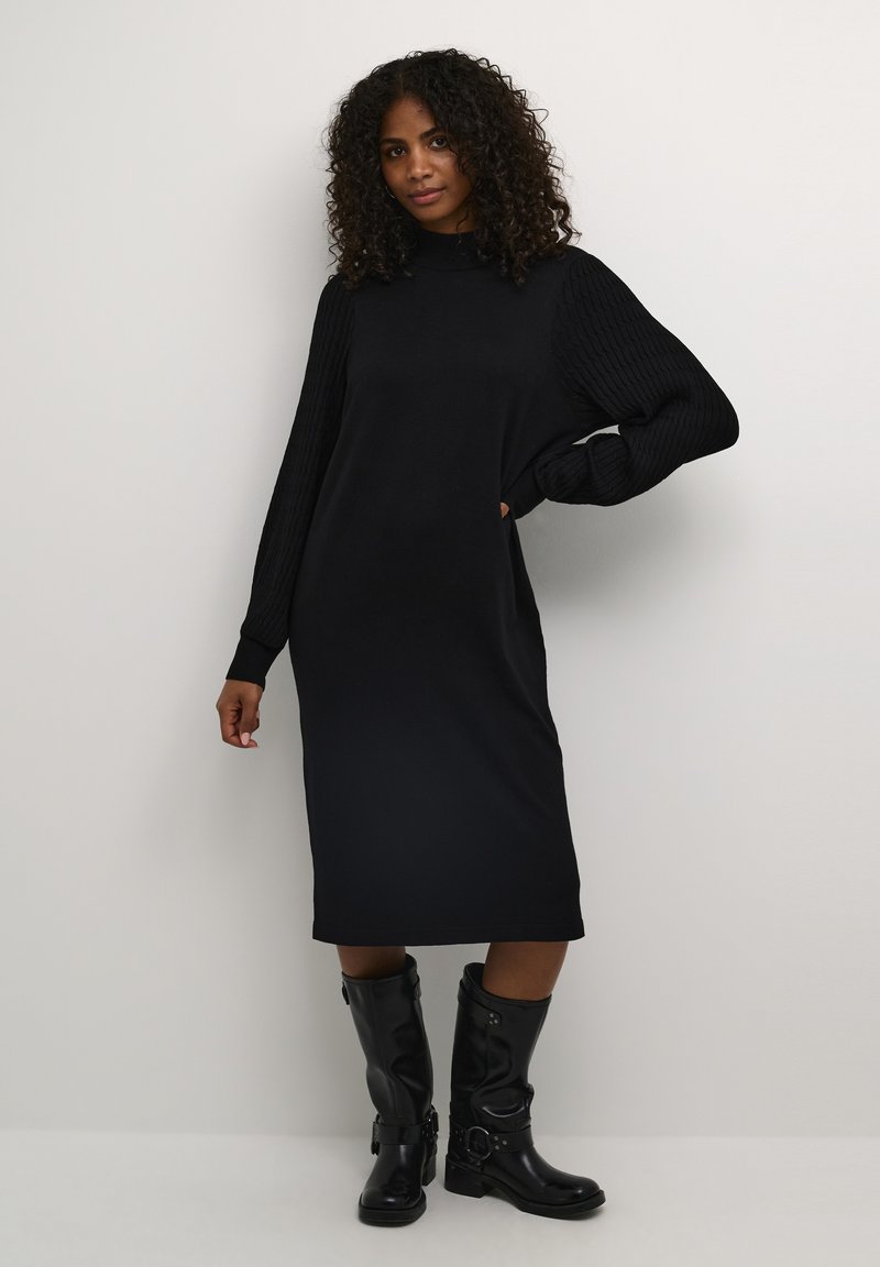 Robe pull noire avec des manches côtelées, longueur genoux, coupe ample. Portée avec des bottes noires jusqu'aux genoux avec des détails en boucle. Texture lisse.