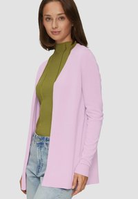Lichtroze open-front cardigan met lange mouwen, gestructureerde gebreide stof en geribbelde manchetten. Gegoed over een groene hoge nek top en blauwe jeans.