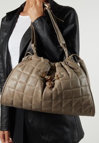 Quiltet beige håndtaske med justerbare skulderremme, detaljer i guldhardware og en samlet top. Glat tekstur med dekorativ syning.