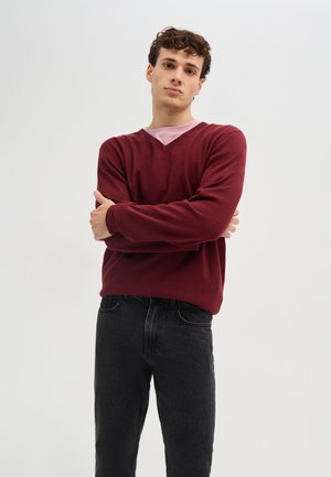 Giovane uomo con capelli ricci che indossa un maglione bordeaux e jeans neri, in piedi con le braccia incrociate davanti a uno sfondo chiaro e semplice.