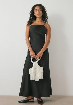 Black satin maxi dress with spaghetti straps, featuring a flowy silhouette. Accessorized with a white, fringed handbag. Black flat shoes.