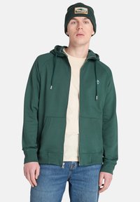Felpa verde con zip, con lacci, tasche frontali e un logo. Indossata sopra una maglietta beige, abbinata a jeans blu e a un berretto coordinato.
