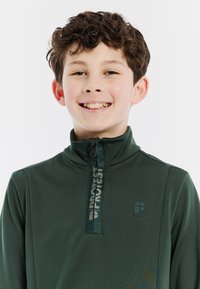 Maglia verde a maniche lunghe con zip e colletto alto. Presenta un logo sul petto e una trama testurizzata sul tessuto. La zip arriva fino al collo.