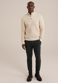 Pulover crem din tricot cu guler cu jumătate de buton, având modele texturate și dungi. Asortat cu pantaloni verzi închis și pantofi maronii.