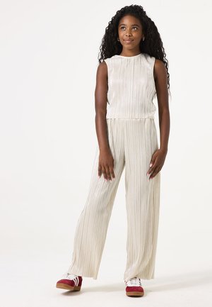 Luxe geplooide jumpsuit in lichtbeige met een hoge halslijn, zonder mouwen en wijde benen. G gedragen met rode en witte sport schoenen.