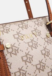 Beige og brun mønstret DKNY håndtaske med læderstropper og guldmetal logo og nitter vist i nærbillede.