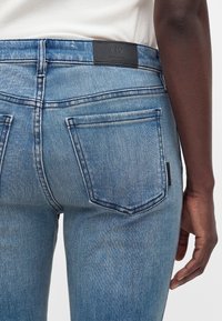 Κοντινό πλάνο ενός ατόμου που φοράει ανοιχτά μπλε τζιν με πίσω τσέπες και μαύρη ετικέτα Armani Exchange στη μέση.